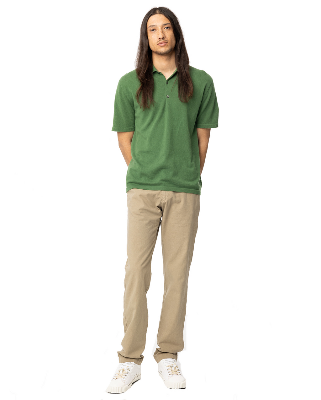 man standing in green polo tee with khaki pants with his hands behind his back. Link leads to men's tee collection
